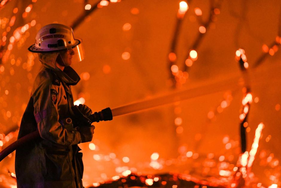 The silhouette of a firefighter is seen in front of a large bushfire burning high into trees.