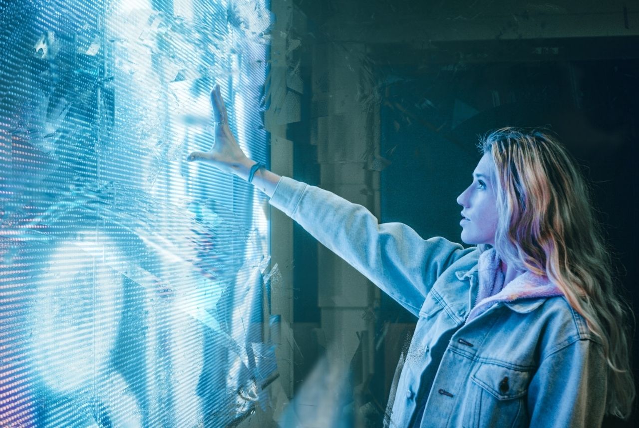woman with long blonde hair wearing a jean jacket holding her hand up to a large blue and white digital screen with abstract 3d graphics on it